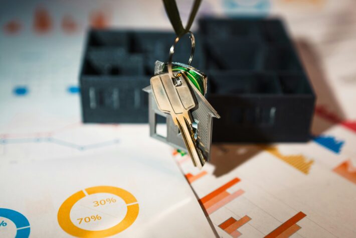 House keys with a miniature house charm hanging above financial charts and graphs, representing the process of finding investment property.