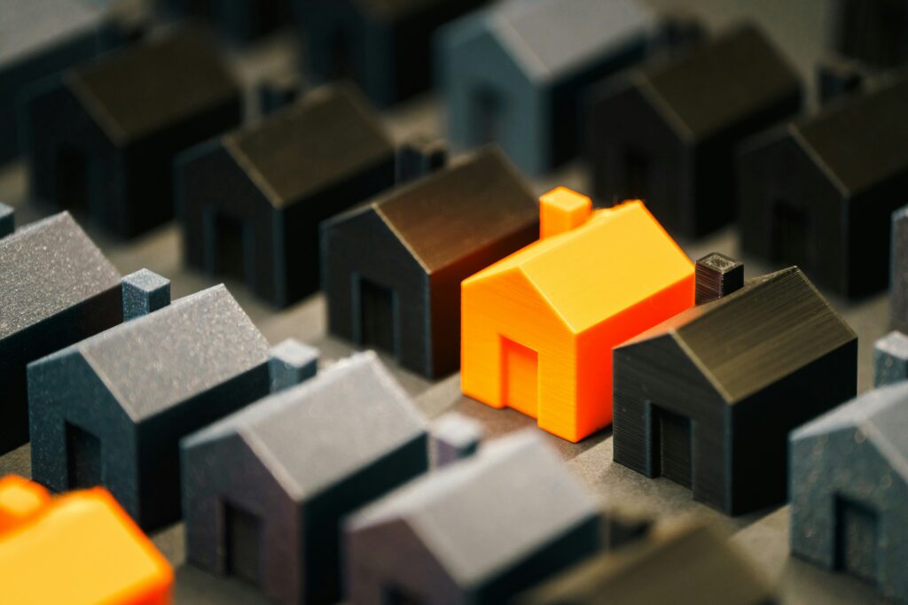 A single orange miniature house stands out among many grey and black houses, symbolizing unique opportunities and real estate market insights.